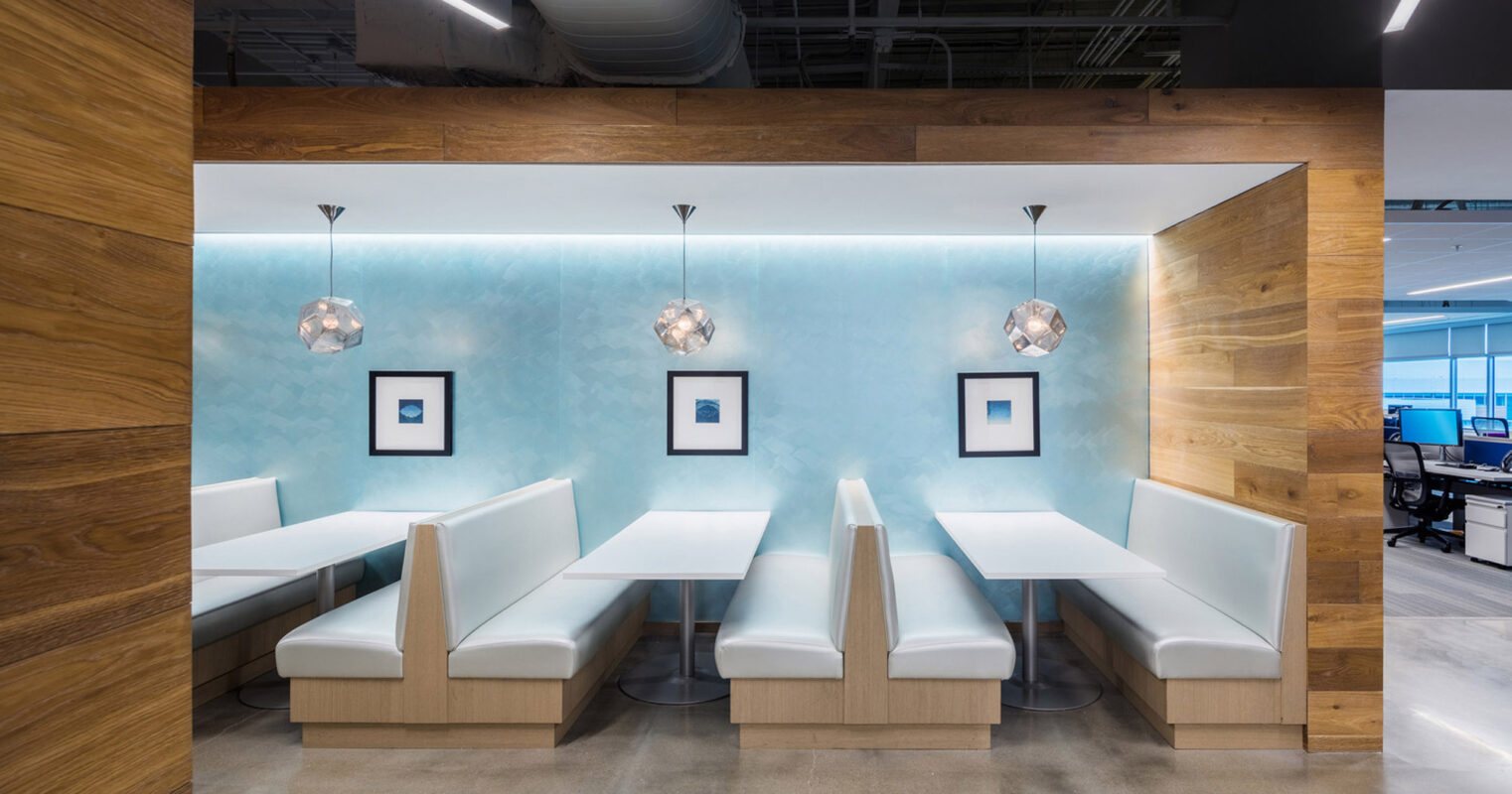 Modern office break room featuring sleek white benches, blue backlit walls, warm wooden paneling, and geometric pendant lighting, creating a fusion of coziness and contemporary design.