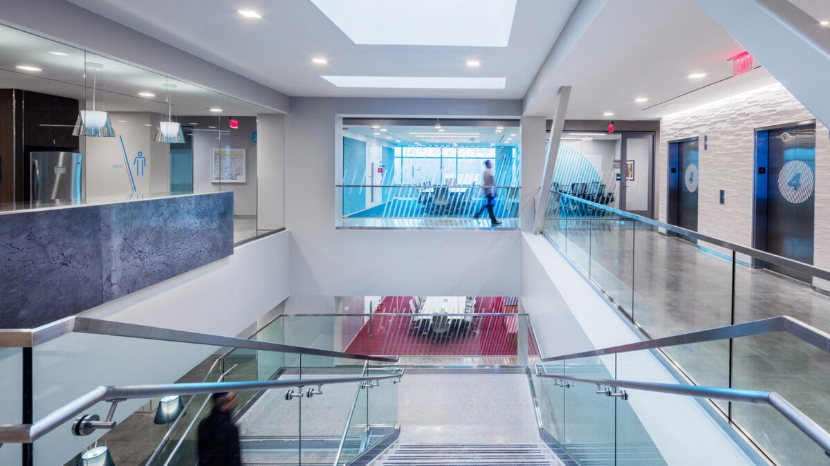 Modern office lobby with glass partitions, sleek metal railing, and dynamic angled white walls that contrast with a vibrant blue accent and reflective flooring. Visible stairway leads to additional levels, enhancing the sense of openness and corporate sophistication.