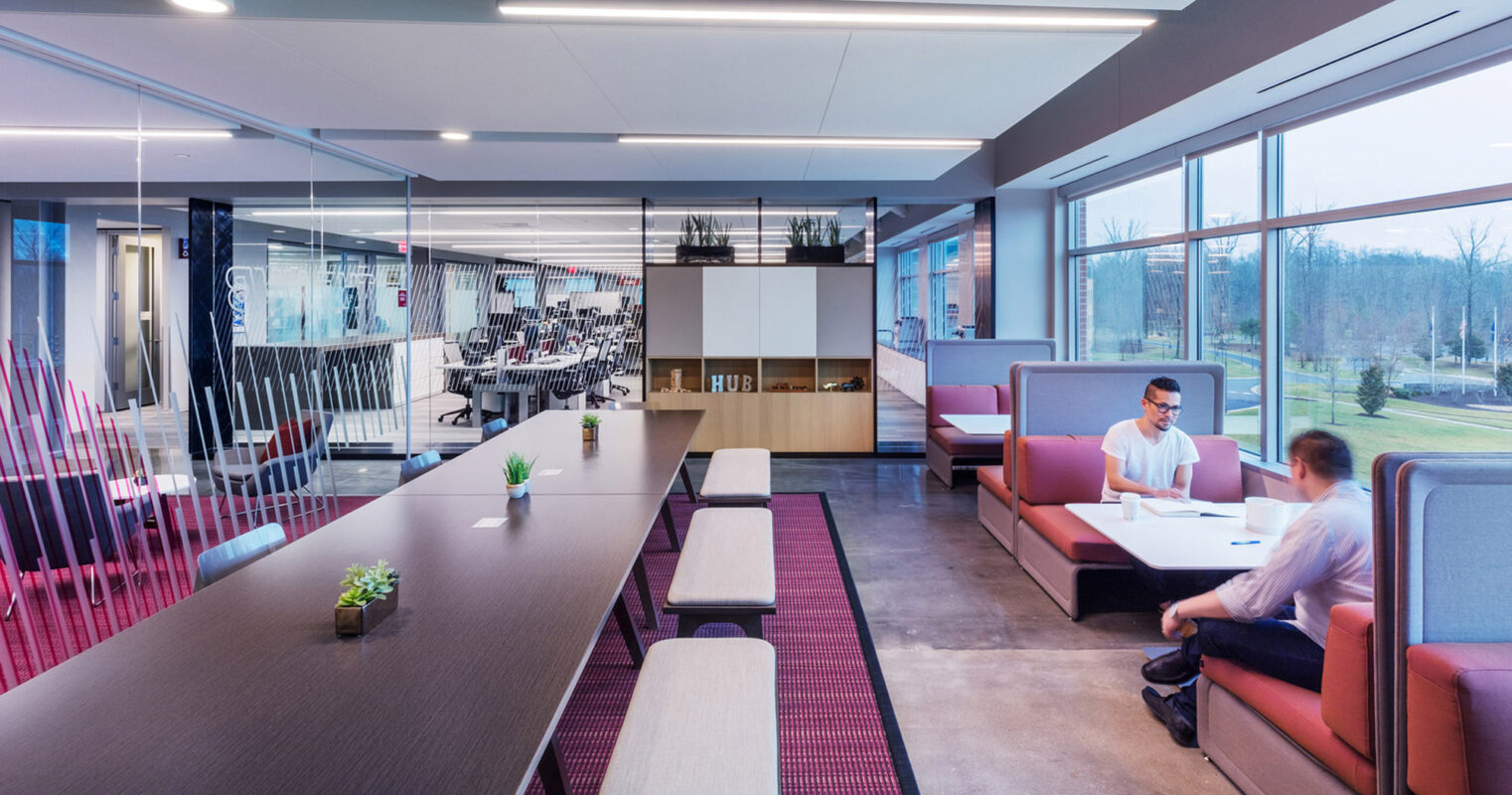 Open-plan office with ample natural light featuring modern furniture, including communal desks and private booth seating with geometric acoustical panels for sound absorption. The color scheme blends neutral tones with vibrant seating upholstery, enhancing the space's dynamic and collaborative atmosphere.
