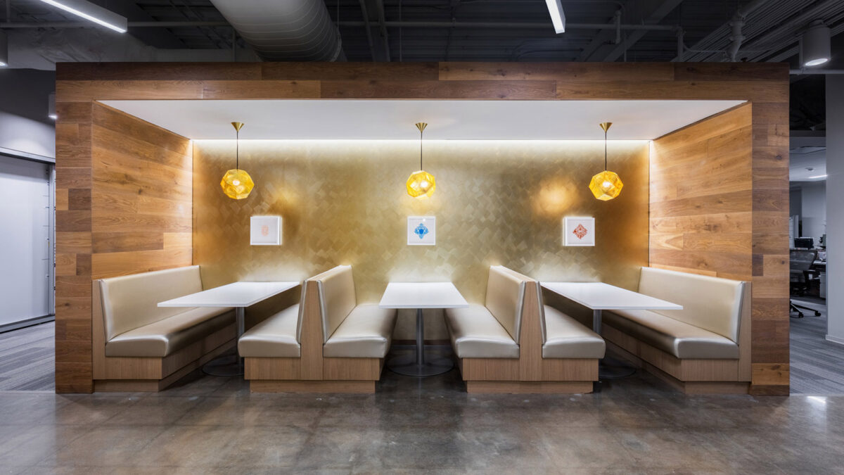 Modern office breakout area featuring warm wood paneling with integrated lighting, complemented by gold textured wallpaper. Three pendant lamps cast a soft glow over two beige banquettes flanking a minimalist white table, creating a cozy yet functional space.