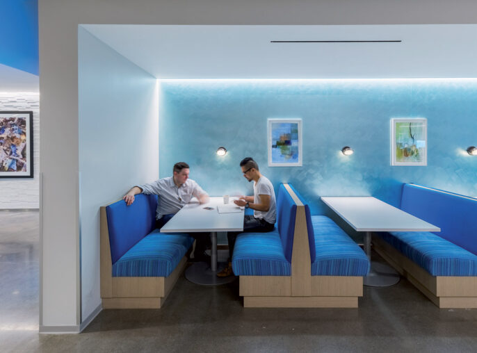 Modern office breakout area featuring vibrant blue upholstered booths, white surfaces, and frosted glass partitions. Strategic lighting accents the space while framed artwork adds a creative touch.