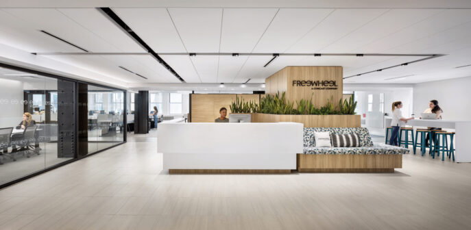 Sleek, modern office foyer featuring a minimalist white reception counter. Behind it, a wood-paneled wall emblazoned with 'FreeWheel' branding anchors the space, complemented by a casual seating area with patterned cushions and a high communal table where employees engage in conversation.