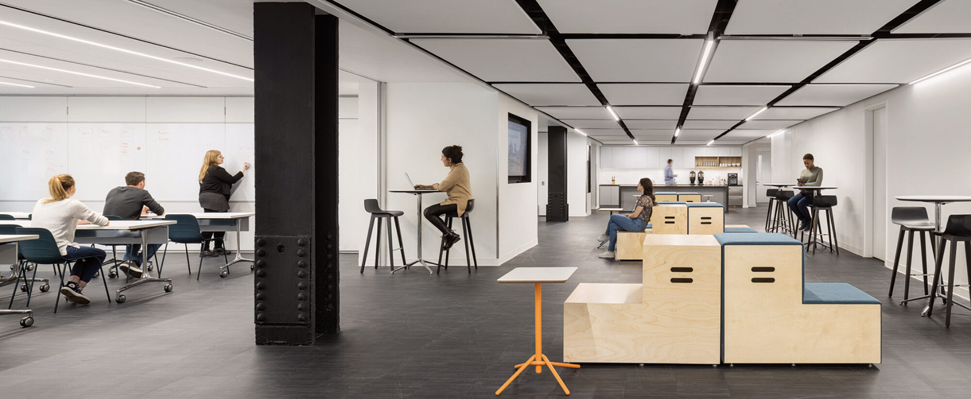 Modern office space with an open floor plan featuring a central plywood staircase, minimalist furniture, and uniform lighting. Strategic seating areas encourage collaboration, while the color contrast adds vibrancy to the monochrome palette.