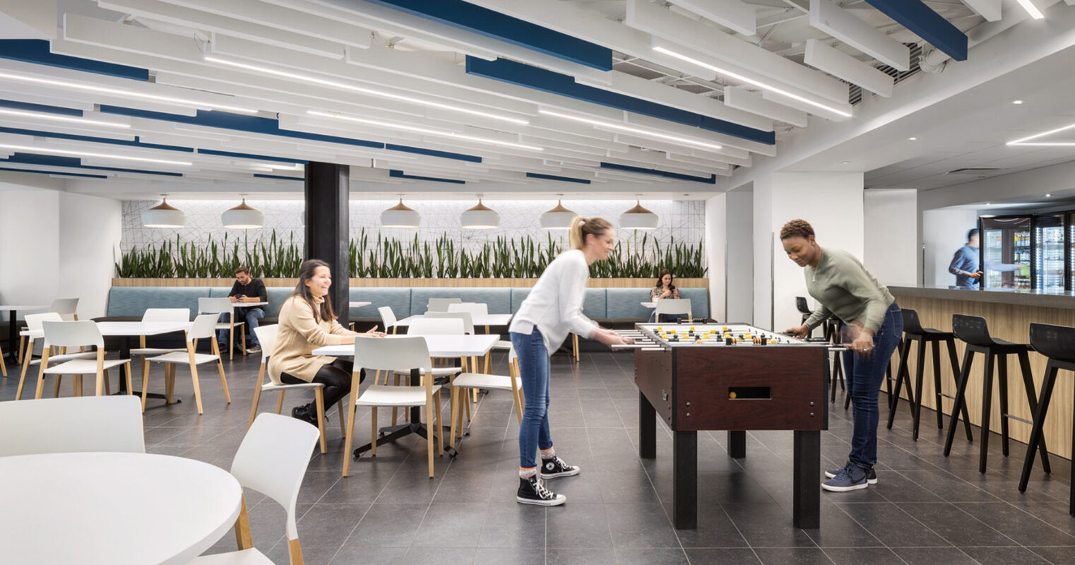 Open-plan office recreation area with contemporary aesthetics, featuring streamlined white tables, high stools, and suspended linear lighting. Two individuals engage in a game of foosball, augmenting the space's casual and collaborative atmosphere. Floor-to-ceiling windows invite natural light, enhancing the plants' vibrant green against the neutral palette.