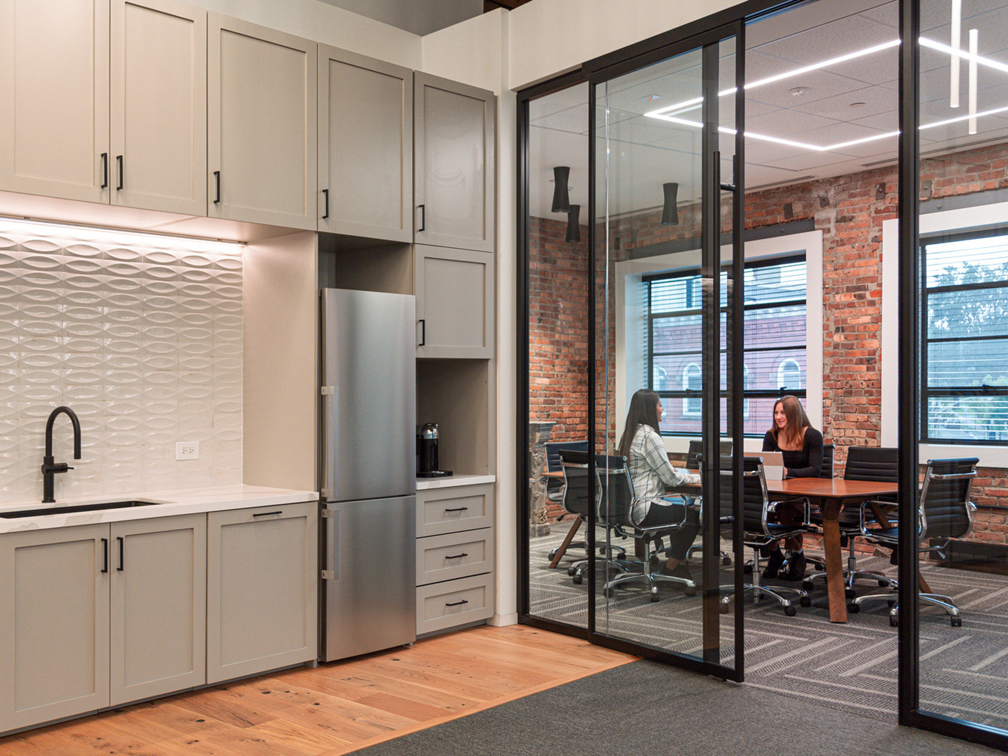 Modern office kitchenette with sleek gray cabinetry and textured white backsplash juxtaposed with warm wood flooring. Glass partition reveals a collaborative workspace with two individuals engaged in conversation. Brick walls add rustic charm, balancing the industrial and contemporary design elements.
