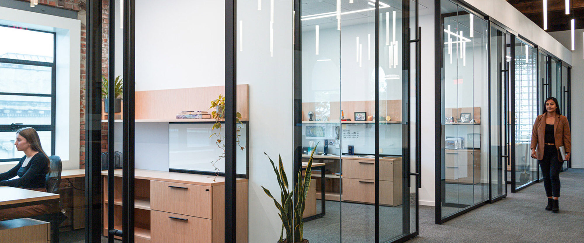 Modern office space highlighting floor-to-ceiling glass partitions, sleek black frames, and wooden desks that create an open yet compartmentalized work environment. Exposed ceiling beams add an industrial touch, while natural light and plants bring warmth to the setting.