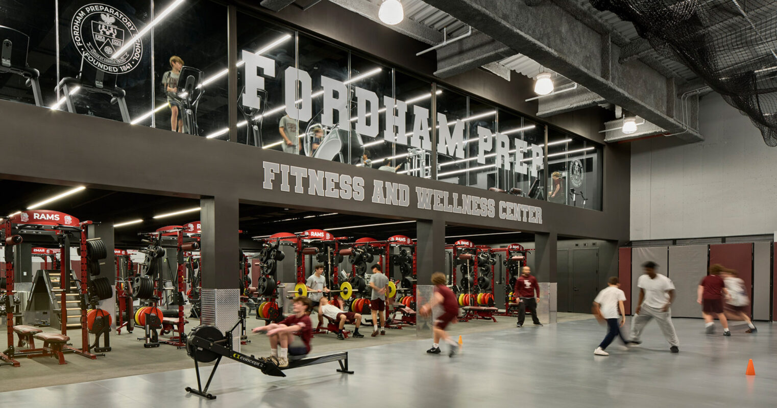 Spacious gym interior with exposed ductwork overhead, reflecting an industrial design. Mirrored walls amplify the space, featuring branded graphics for Fordham University. Red and black gym equipment are organized for functional training, promoting a vibrant, energetic atmosphere.