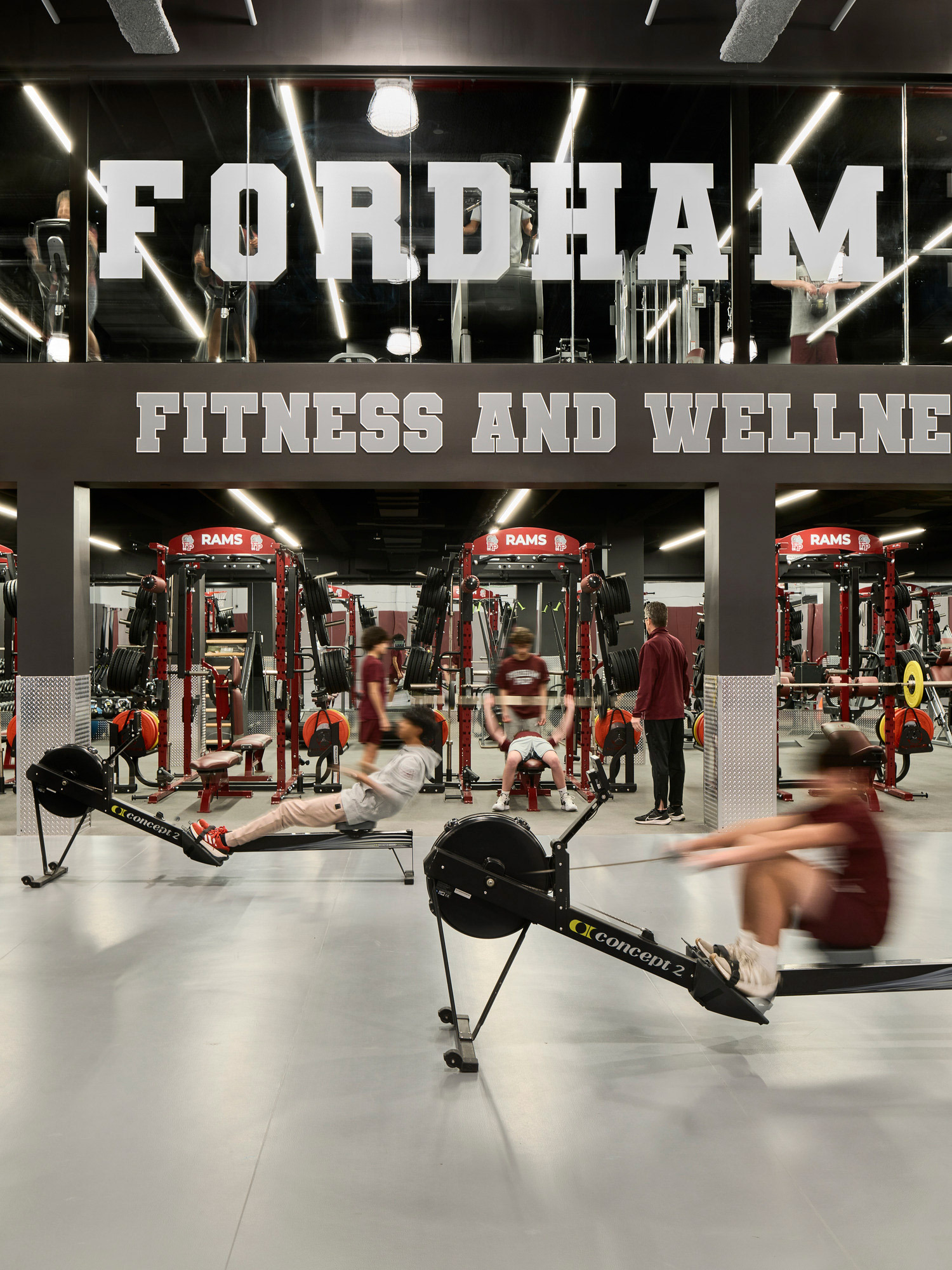 Modern gym interior with black walls, red and gray fitness equipment, and large bold 'FORDHAM FITNESS AND WELLNESS' lettering on mirror-finished ceiling above. People are actively using rowing machines and strength-training apparatus, indicating a dynamic and well-facilitated workout environment.