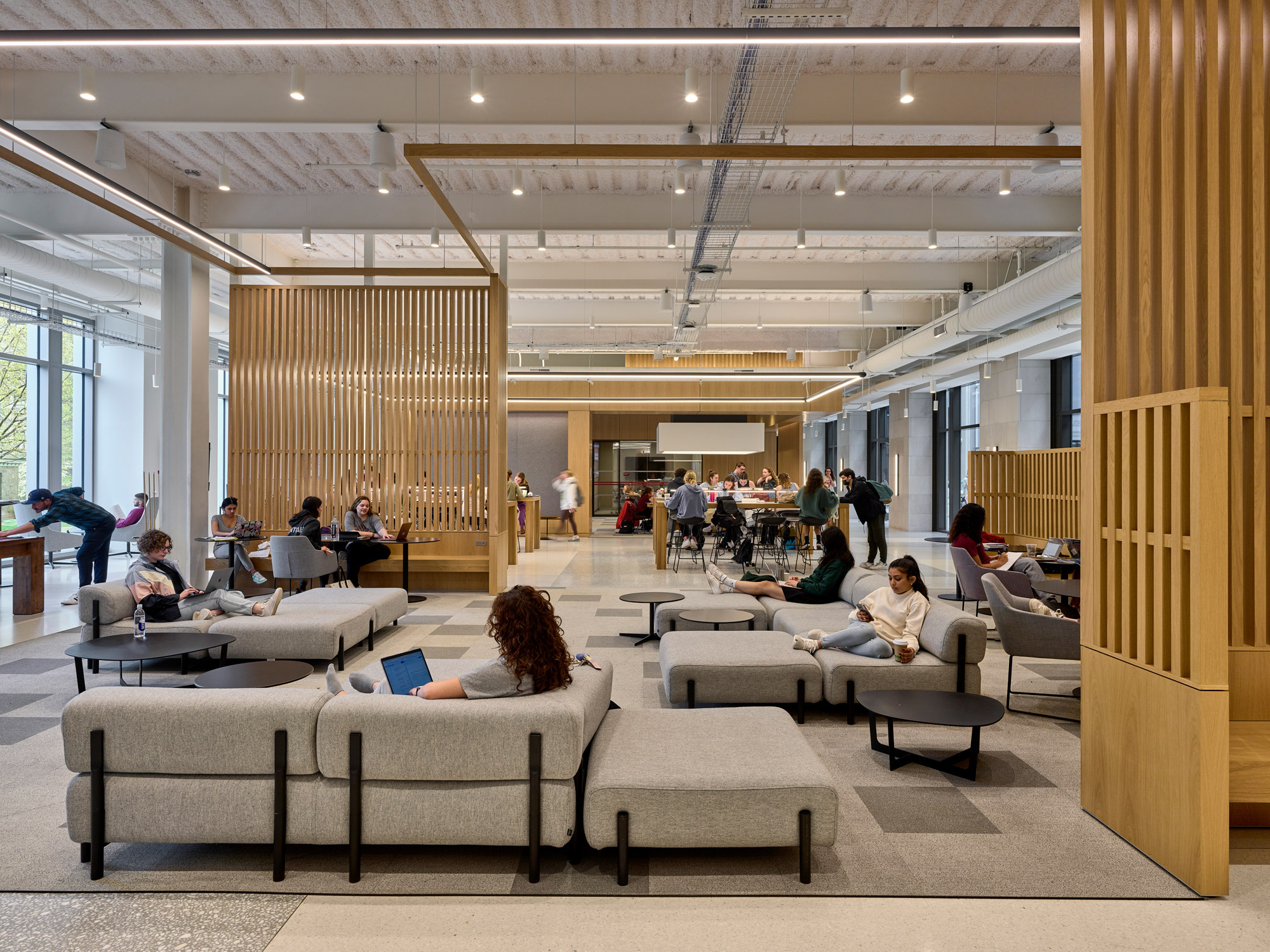 Open-plan office space featuring modular gray seating, slatted wood partitions, and a high exposed ceiling. Natural light filters through large windows, highlighting the warm tones of the wooden accents and the neutral color palette. People are working and interacting in a contemporary collaborative environment.