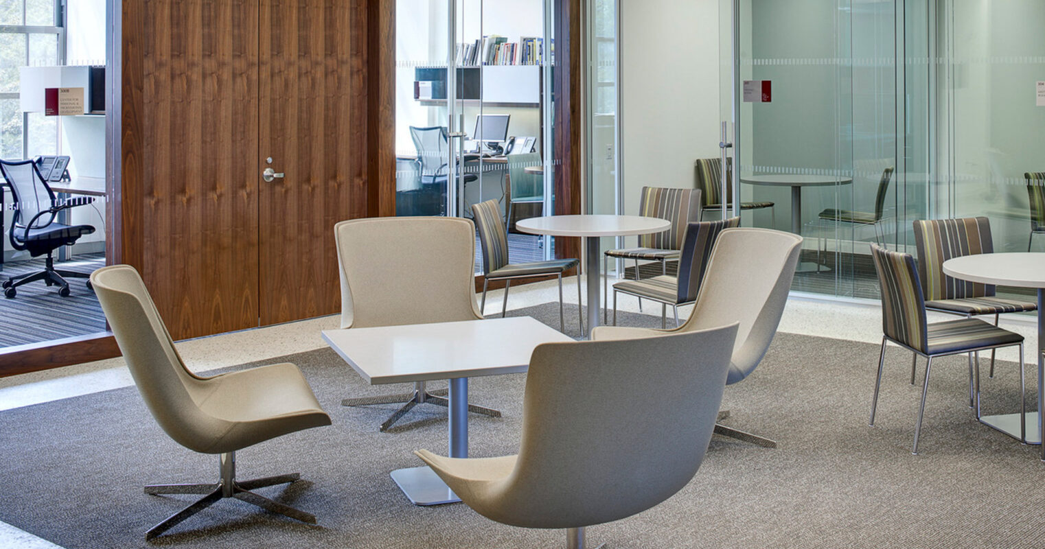 Modern office space featuring neutral tones with sleek, taupe armchairs, a textured gray area rug, wood grain doors, and frosted glass partitions promoting a light, airy atmosphere conducive to collaboration.