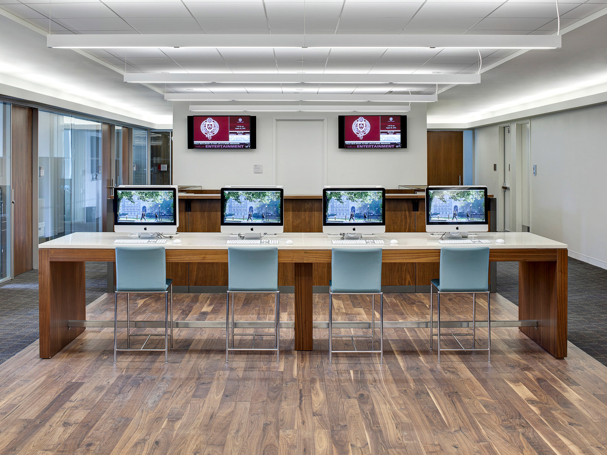 Contemporary office interior featuring a cluster of four desk stations with teal chairs, set against a wood floor, flanked by dual flat-screen monitors and underlined by sleek, recessed lighting in a drop ceiling design. A logo-emblazoned wall serves as the backdrop.