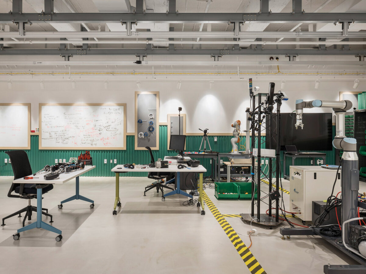 Modern industrial-style workspace featuring exposed ductwork and concrete floors. Central tables with electronic equipment suggest a collaborative tech lab environment, complemented by wall-mounted whiteboards adorned with diagrams, enhancing the space's creative and analytical functionality.