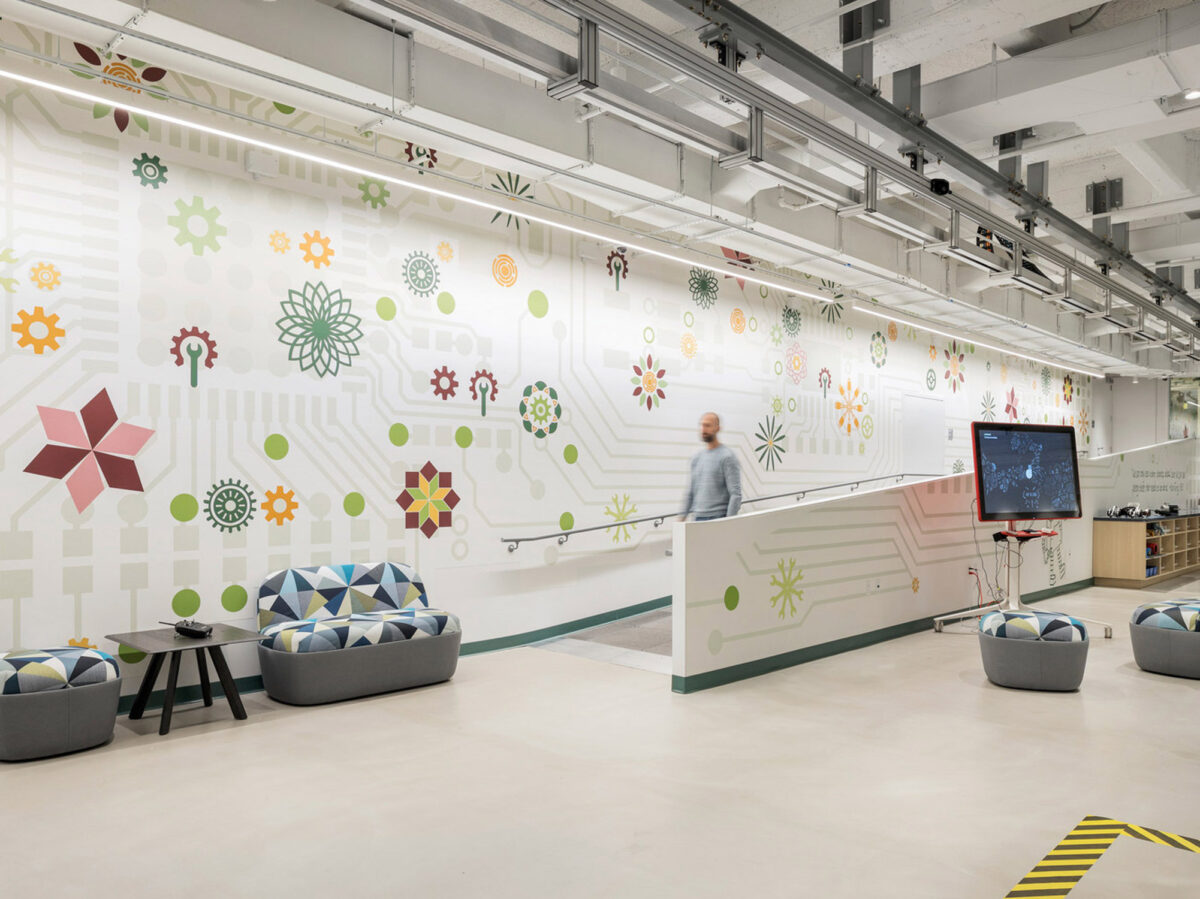 Contemporary office breakout area featuring an eclectic mix of geometric and organic patterns on a white wall, accented with colorful graphics. The space includes cushioned poufs centered around a mobile display screen, under exposed ceiling utilities for an industrial feel.