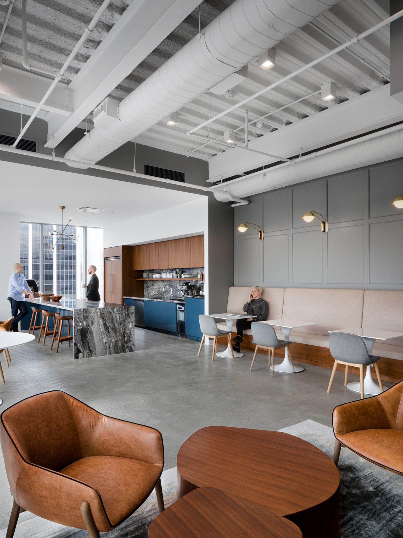 Modern office break room blending industrial and contemporary styles, featuring exposed ceilings, pendant lighting, and a marble kitchen island. Comfortable seating areas with wood tables and leather chairs provide a casual workspace. Employees engage in relaxed conversations.