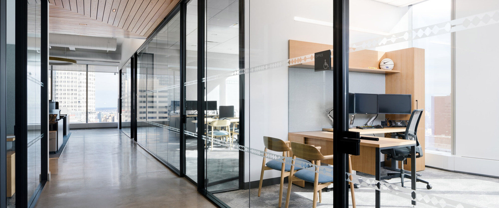 Spacious modern office interior featuring glass partitions, a reflective concrete floor, and a warm wooden slat ceiling that contrasts the cool-tone walls, populated with ergonomic chairs and sleek workstations under natural daylight.