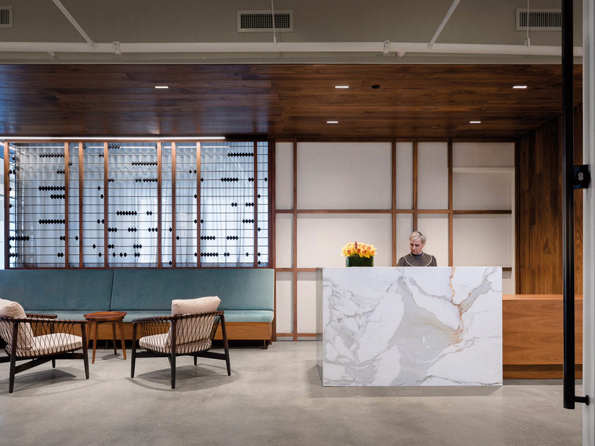 Reception area with minimalist design, featuring a marble-front desk, warm wooden ceiling beams, and tailored upholstered seating. Frosted glass partitions and pendant lighting add to the refined ambiance.