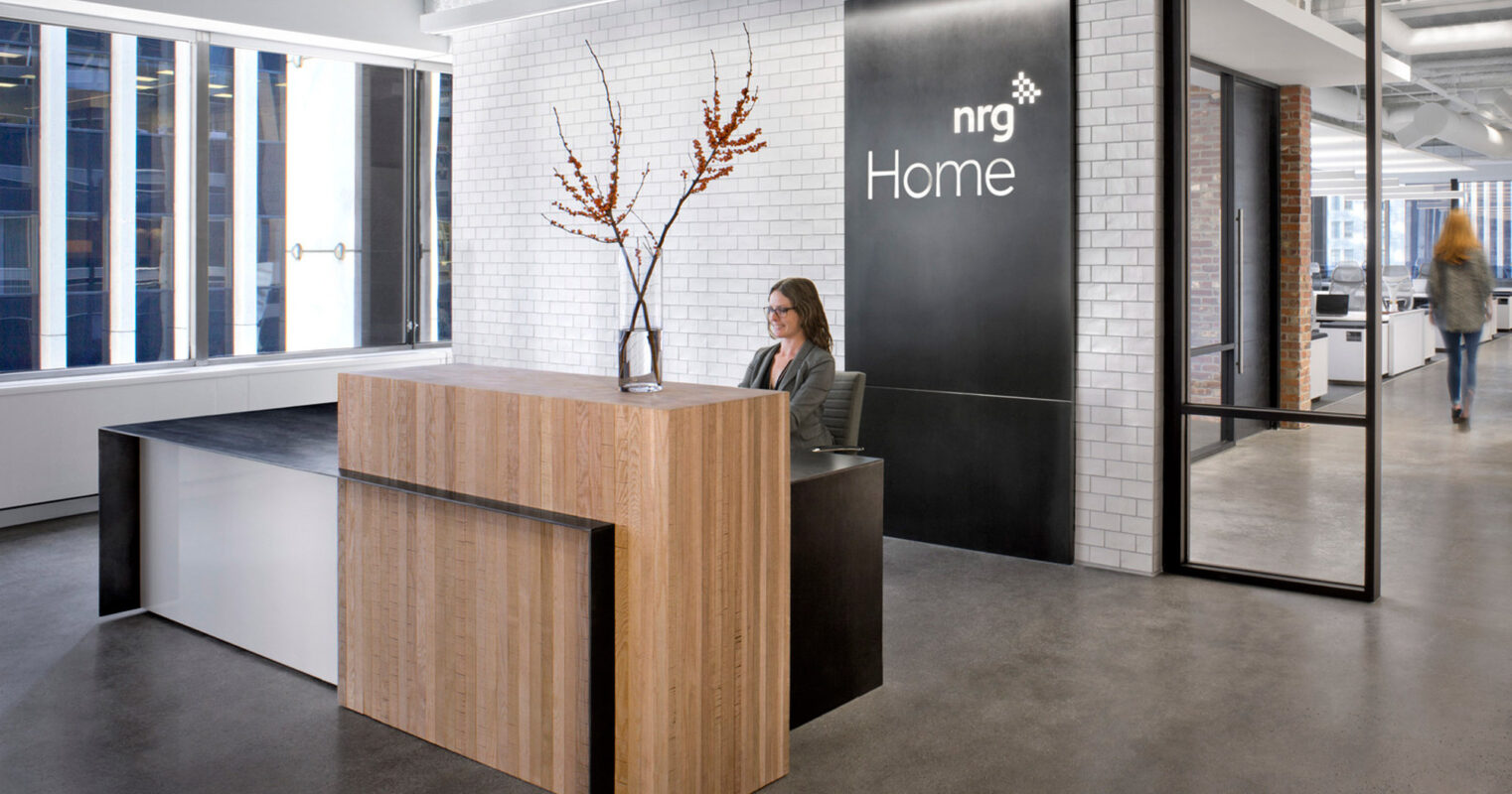 Modern, minimalist reception area featuring a sleek, wooden desk with a clear vase of tall branches. The design incorporates ample natural light, clean lines, and a neutral color palette, complemented by industrial-style doors and concrete flooring.