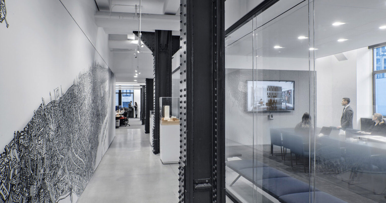 A spacious modern office corridor with industrial-style exposed ceiling piping, contrasting with sleek glass partition walls and monochromatic art. Staff navigate the open-plan workspace beyond, visible through the transparent interfaces that enhance a sense of connectivity and light flow.
