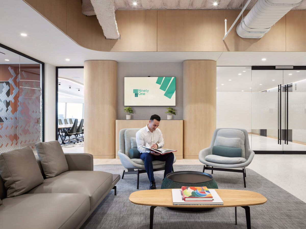 Modern office lobby with sleek furnishings and a neutral color palette. Natural light enhances the warm wooden reception desk, flanked by plush seating and contemporary art pieces. Visible glass partitions reveal a collaborative workspace beyond.