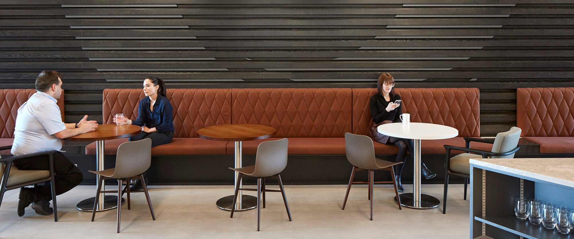 Modern café interior featuring terracotta-hued upholstered booths, circular wooden tables, and minimalist chairs. A striking black textured accent wall contrasts with the warm tones, complemented by sleek horizontal lighting elements and an acoustical tile ceiling for a contemporary ambiance.