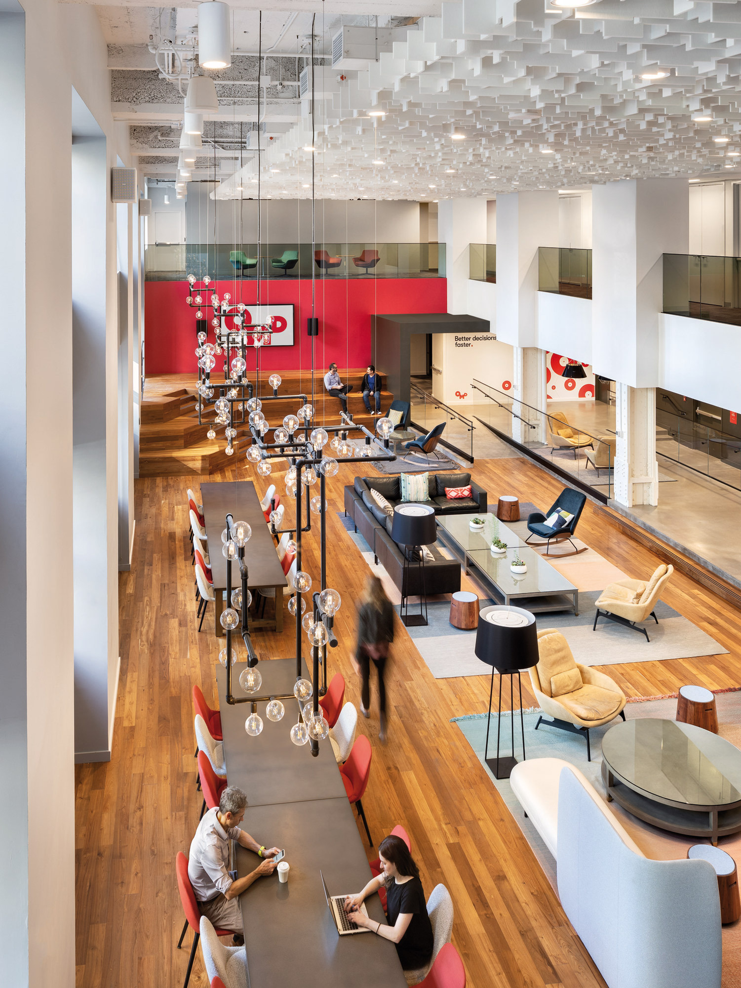 Spacious modern lobby with exposed ceiling, floating white acoustic panels, and pendant lighting. A mix of red accent walls, wooden floors, and contemporary furniture create a dynamic communal space.