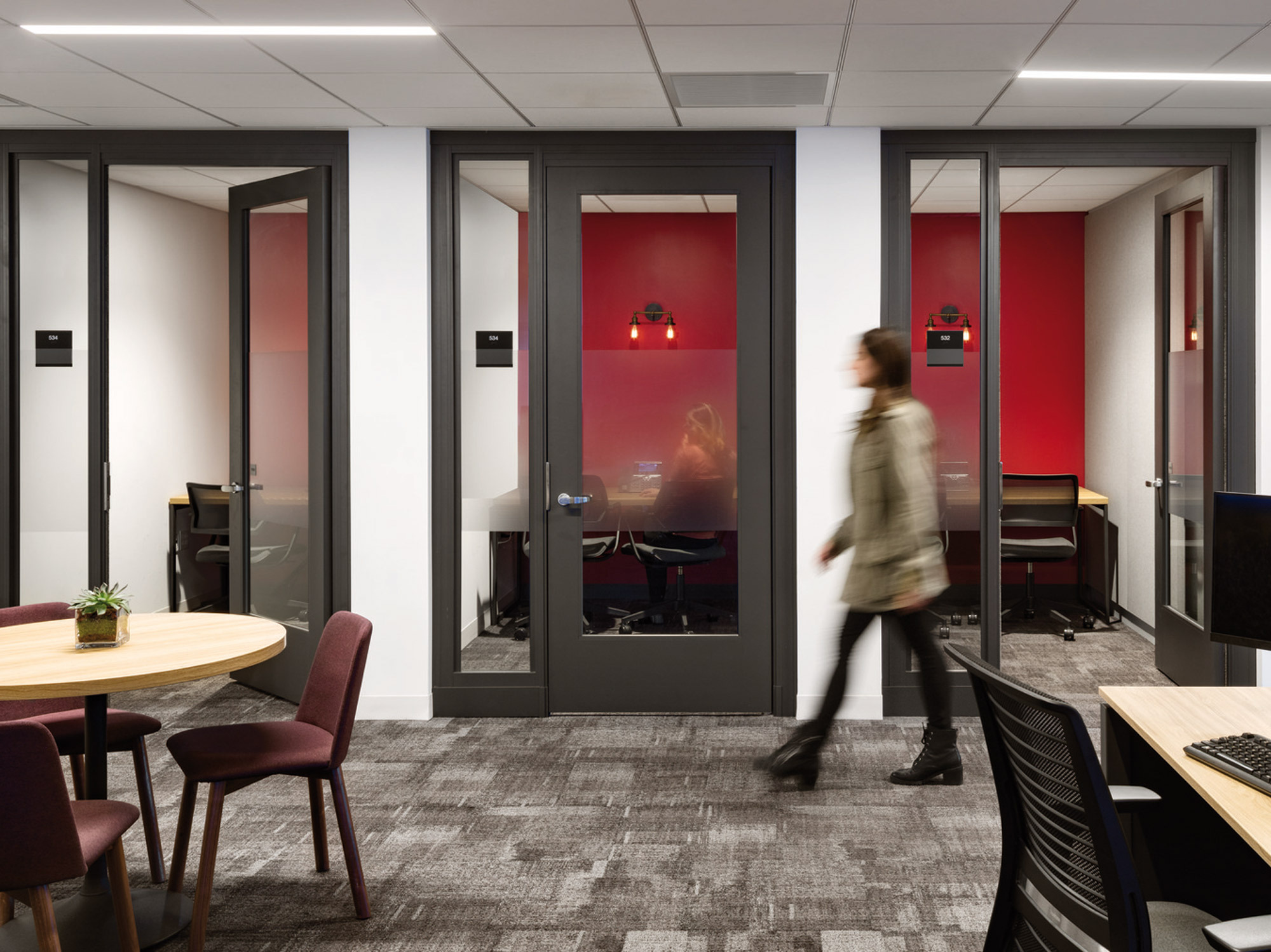 Modern office interior showcasing glass-walled meeting rooms with frosted lower panels for privacy. Red pendant lighting adds a pop of color against neutral tones, while a person in motion suggests a dynamic work environment. Gray carpet and wood accents blend professionalism with comfort.