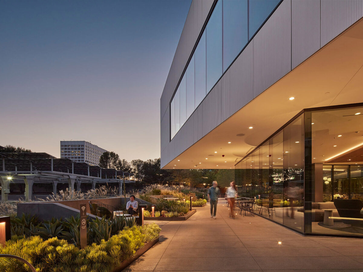 Modern office building at twilight with floor-to-ceiling windows offering a glimpse into the warmly lit interior. The building features sharp geometric lines and a minimalist design, complemented by surrounding landscaped greenery and outdoor seating areas.