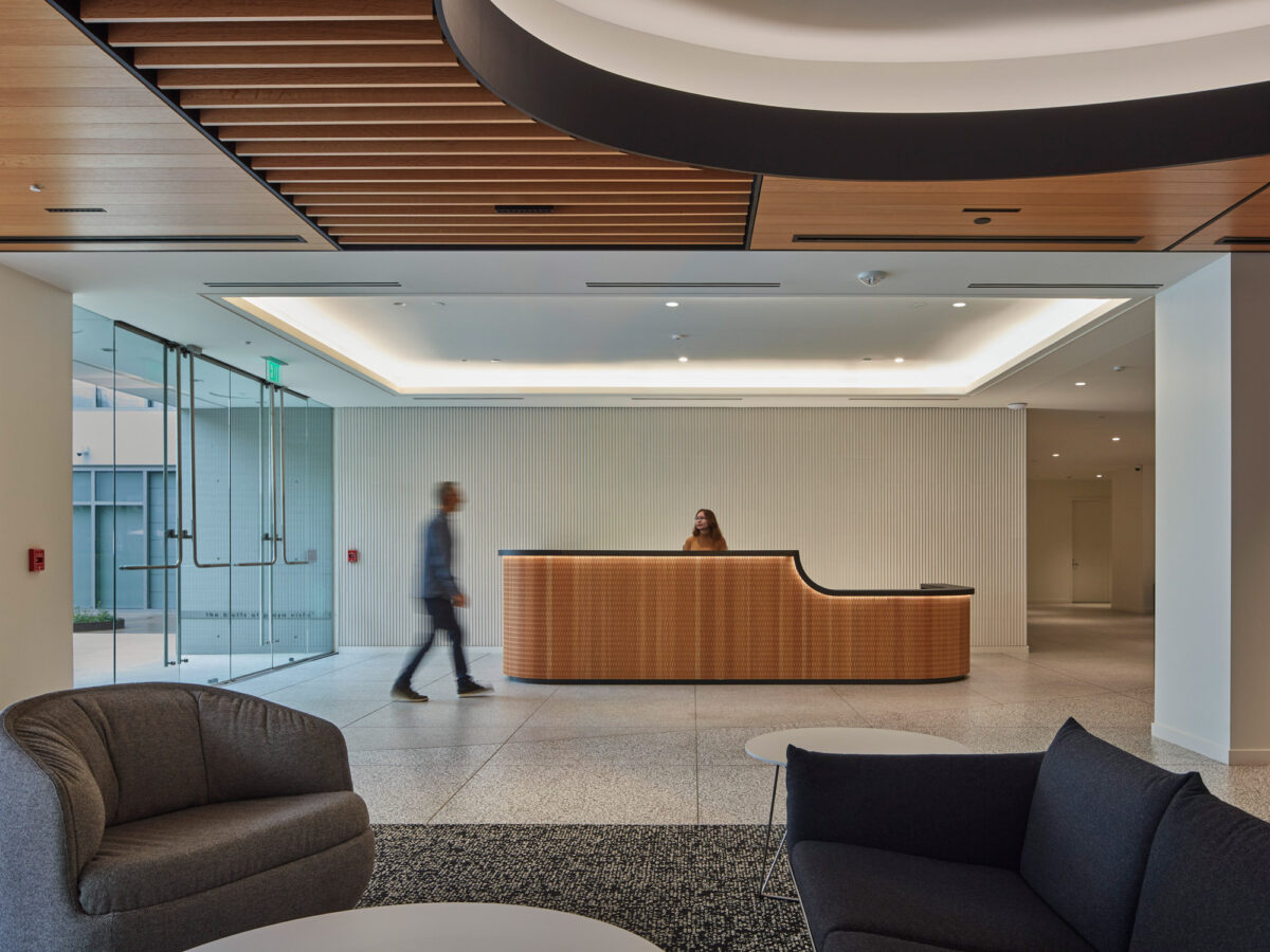 Modern corporate lobby with a curved wooden reception desk, slatted ceiling accents, and sleek white walls. Ambient lighting and plush seating offer a welcoming atmosphere, while a blurred figure conveys motion, suggesting a dynamic business environment.