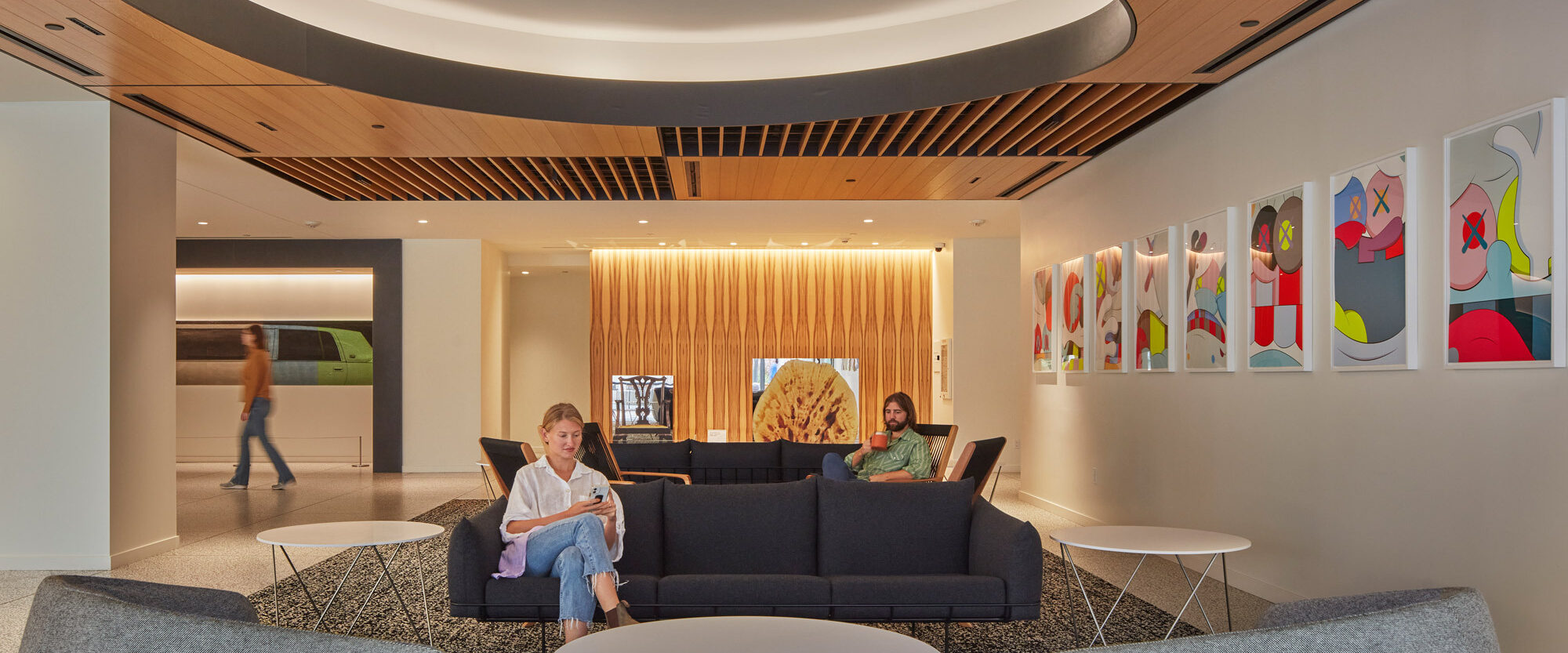 Modern lobby with a concentric circular ceiling feature, wood slat accents, and a central seating area with a sleek sectional sofa, accompanied by a dog and two people engaged in relaxation. Vibrant artwork adds a pop of color to the serene, neutral-toned space.