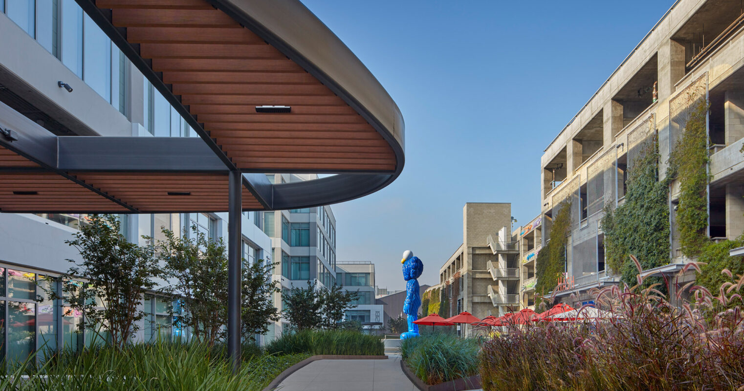 Modern mixed-use development with sweeping curved roofline, incorporating natural wood and concrete with landscaped green walkways, juxtaposed by a dynamic blue sculpture, enhancing the aesthetic and pedestrian experience.
