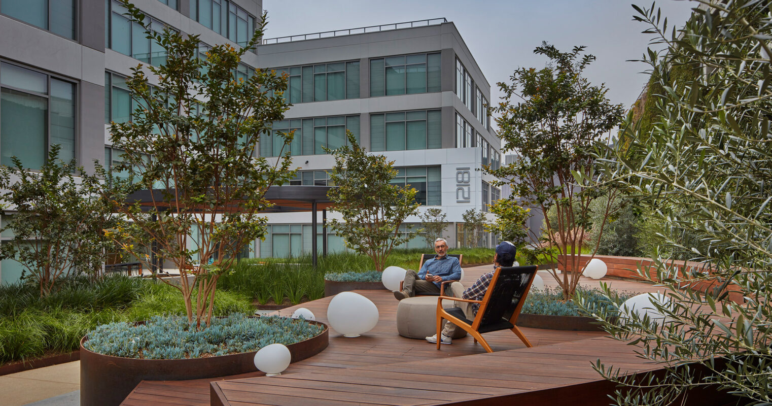 Modern exterior breakout space with an organic design, featuring wooden curved benches interspersed with lush greenery and spherical white lights, complementing the angular architecture of surrounding buildings.