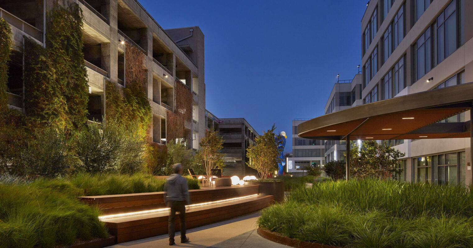 Evening view of a modern urban courtyard with integrated landscaping. Features include stepped wooden planters, ambient lighting, and contemporary outdoor seating. Industrial-style buildings flank the space, enhancing the blend of natural and architectural design.