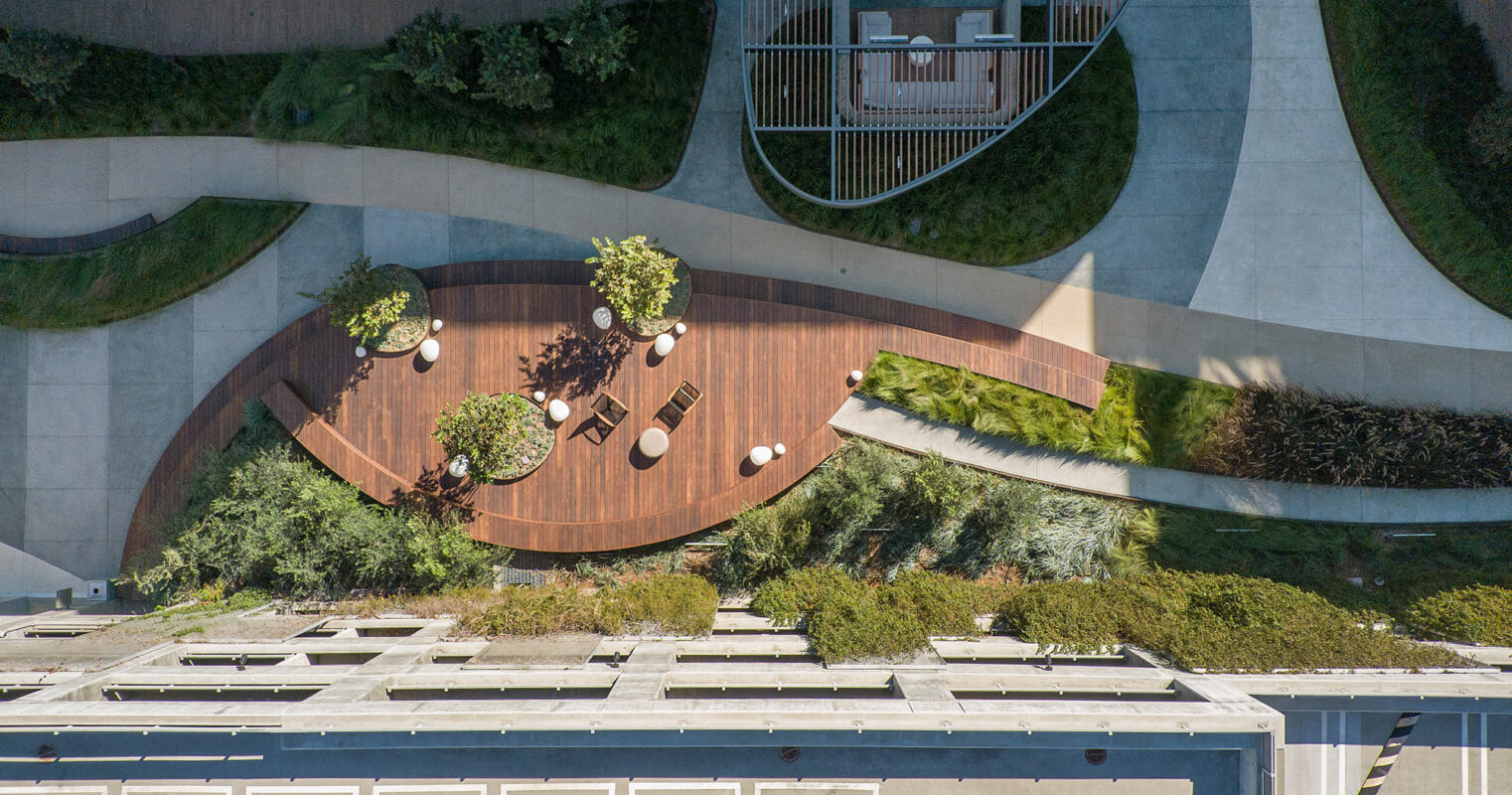 Aerial view of a landscaped rooftop garden, featuring curved wooden pathways, interspersed with green planting areas, and modern outdoor furniture for relaxation. The design seamlessly integrates natural elements with the building's architecture.