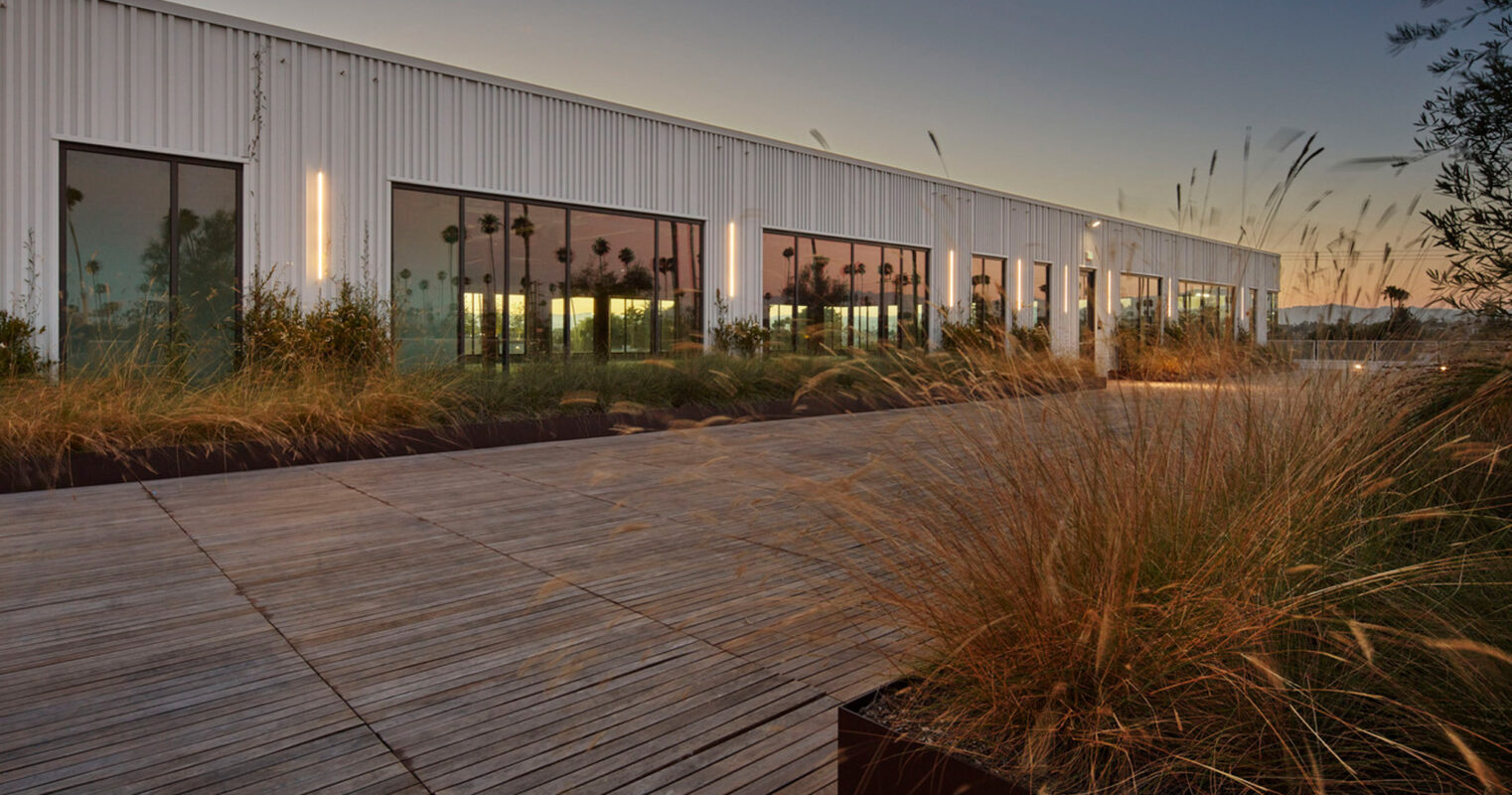 Contemporary single-story building with a reflective metal facade and expansive glass windows at dusk, featuring an elevated wooden deck surrounded by tall grasses in a natural landscape setting.