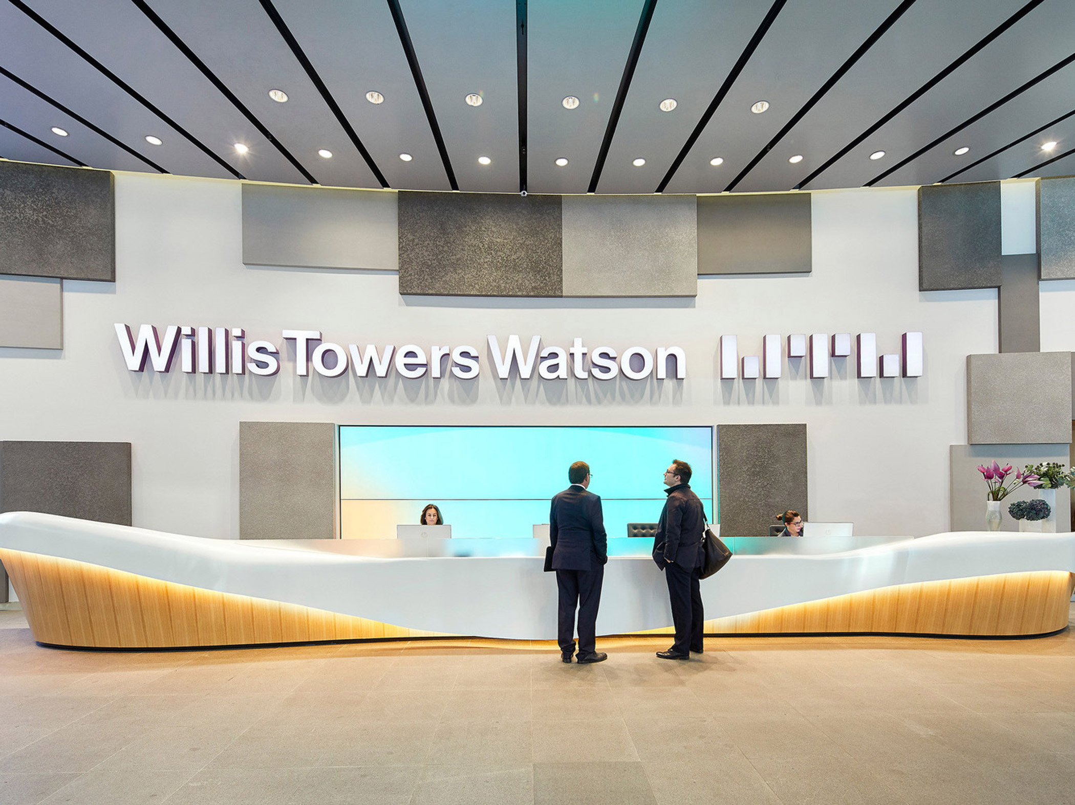 Modern corporate reception area featuring a curved white desk with ambient lighting, flanked by two individuals in business attire. The backdrop showcases the company name, 'Willis Towers Watson,' in large lettering, set against contrasting gray acoustic panels, beneath a sleek strip-lit ceiling.
