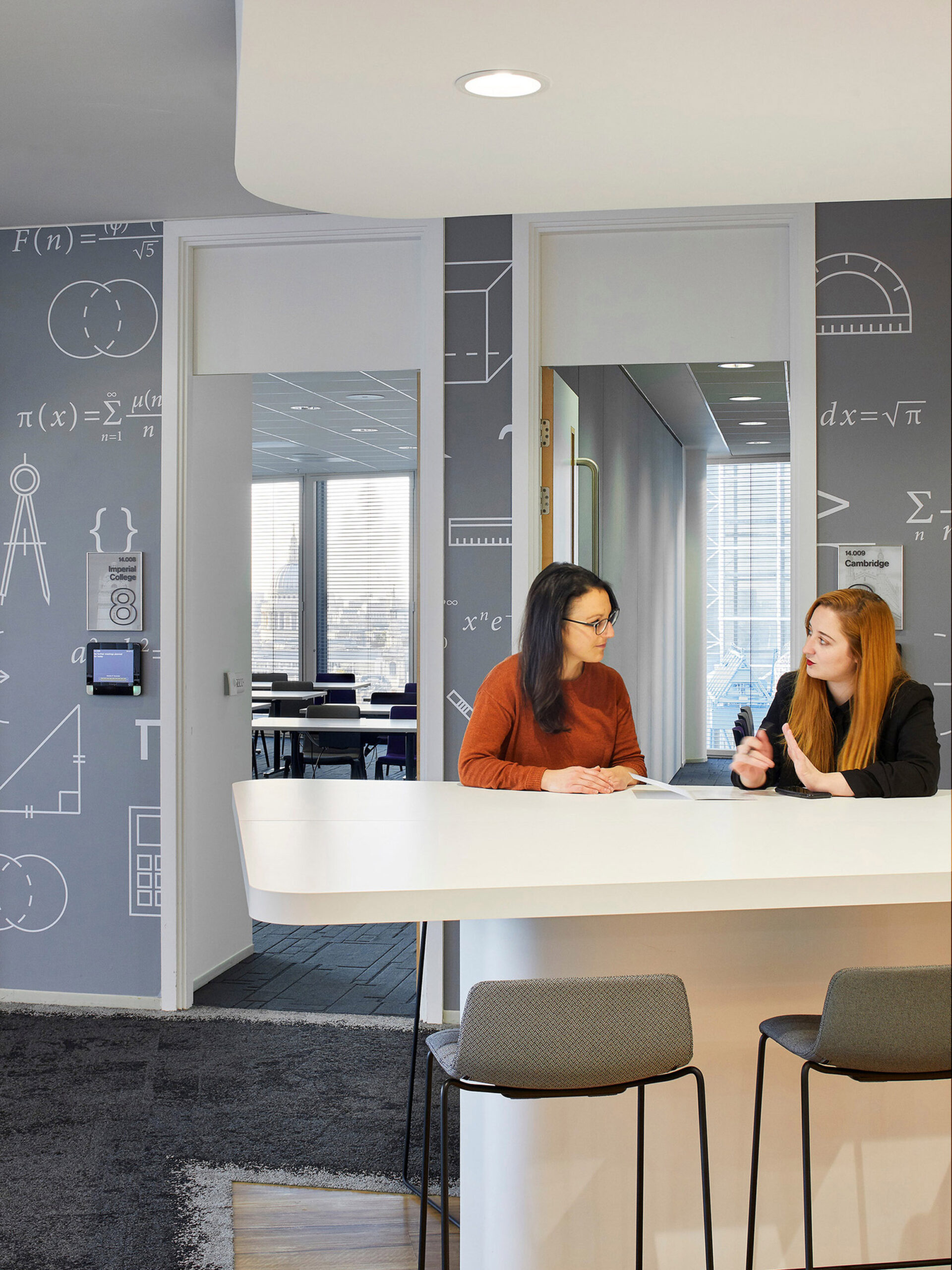 A contemporary office break room, featuring charcoal gray flooring, sleek white cabinetry, and a geometric-patterned accent wall with mathematical illustrations. Two professionals engage in conversation at a minimalist table with functional seating, illuminated by natural light from adjacent windows.