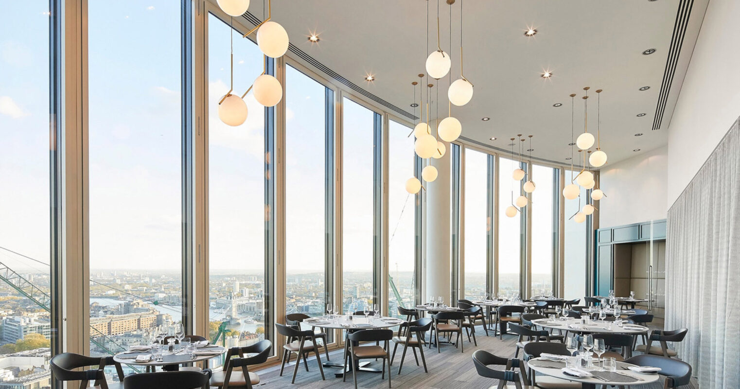 Modern restaurant space with floor-to-ceiling windows offering expansive cityscape views. The interior features sleek minimalist furniture, with a neutral color palette and strategically placed spherical pendant lights creating a rhythm in the vertical space.