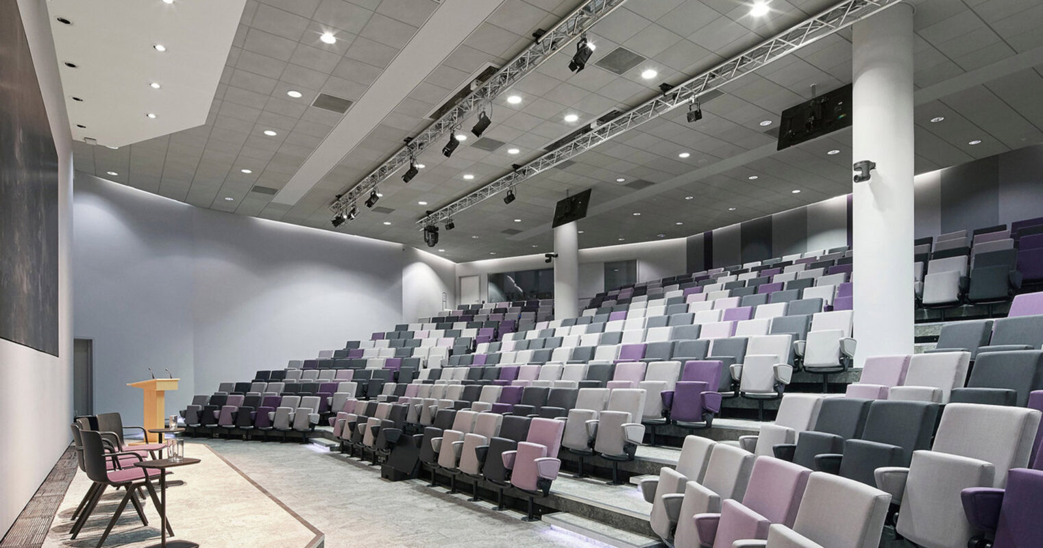 Contemporary auditorium with tiered seating featuring shades of gray and purple upholstered chairs, focused lighting from ceiling spots and ambient wall fixtures, and a central stage with a wooden podium flanked by chairs.