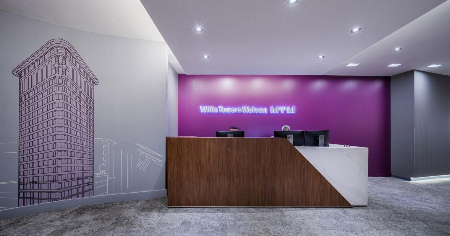 Elegant office lobby featuring a vibrant fuchsia accent wall with Willis Towers Watson branding and a line drawing of a skyscraper, complemented by a sleek, wooden reception desk and modern, recessed ceiling lighting.