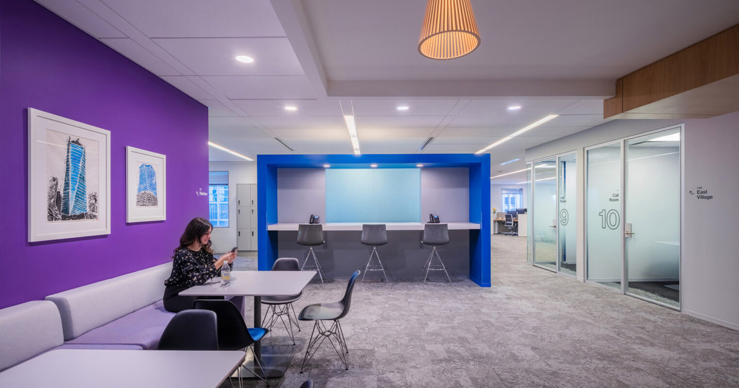 A modern office space featuring a vibrant purple accent wall, sleek furnishings, pendant lighting, framed skyscraper artwork, and a woman working at a white bench-style table. Glass doors and wooden panels enhance the open, collaborative layout.