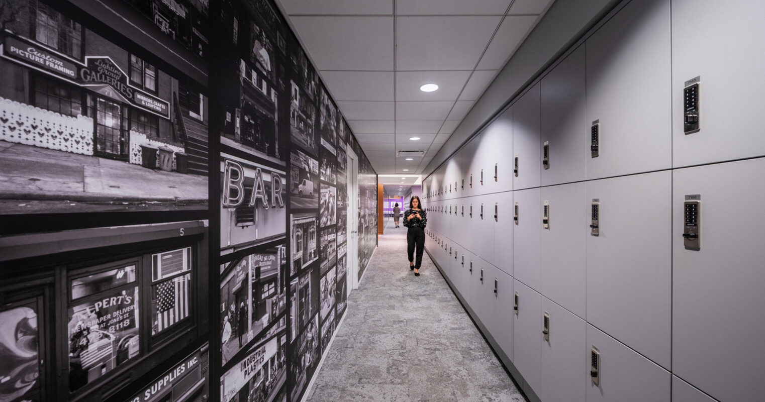 Hallway lined with sleek, white lockers on one side and a large-scale, vintage-style photographic mural on the other, creating a striking contrast. A person walks down the corridor, adding a dynamic element to the scene.