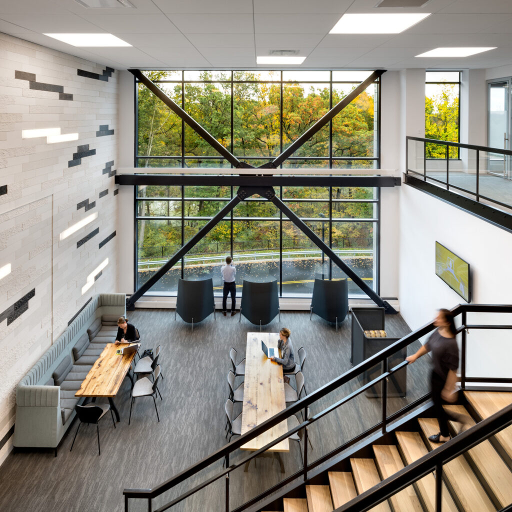 Modern office interior with high ceilings and large picture windows showcasing an autumnal tree panorama. Natural light illuminates the minimalist décor, contrasting the industrial-style black beams and railings. Open-plan seating area features wood tables and gray upholstered benches, fostering a collaborative workspace