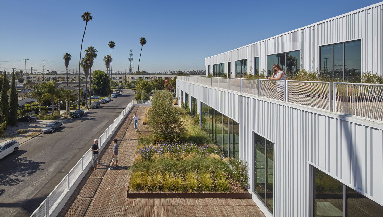 Modern office building integrating green spaces: an employee enjoys a lush rooftop garden walkway, with palm trees and blue skies in the background, emphasizing eco-friendly design and work-life balance.