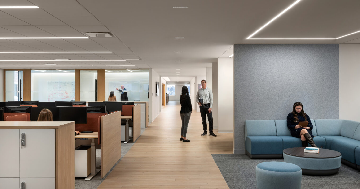 Bright and spacious modern office corridor featuring collaborative spaces. Soft neutral color palette enhances the inviting atmosphere, while strategically placed recessed lighting illuminates the area. Minimalist furniture, including a curved blue sofa and round ottoman, encourages informal meetings and relaxation, promoting a productive work environment.