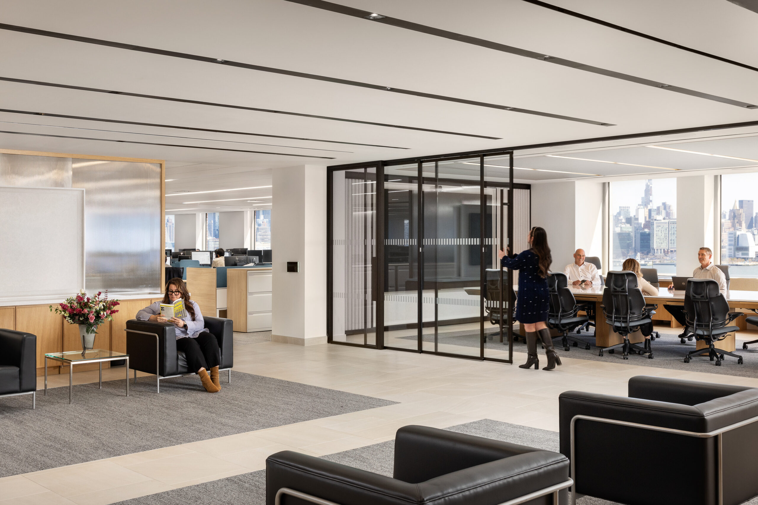 A modern open-plan office lobby features sleek black leather seating and a soft gray area rug, creating a welcoming atmosphere. Large glass partitions delineate a conference room, while natural light floods the space through expansive windows, highlighting the minimalist design and contemporary furnishings.