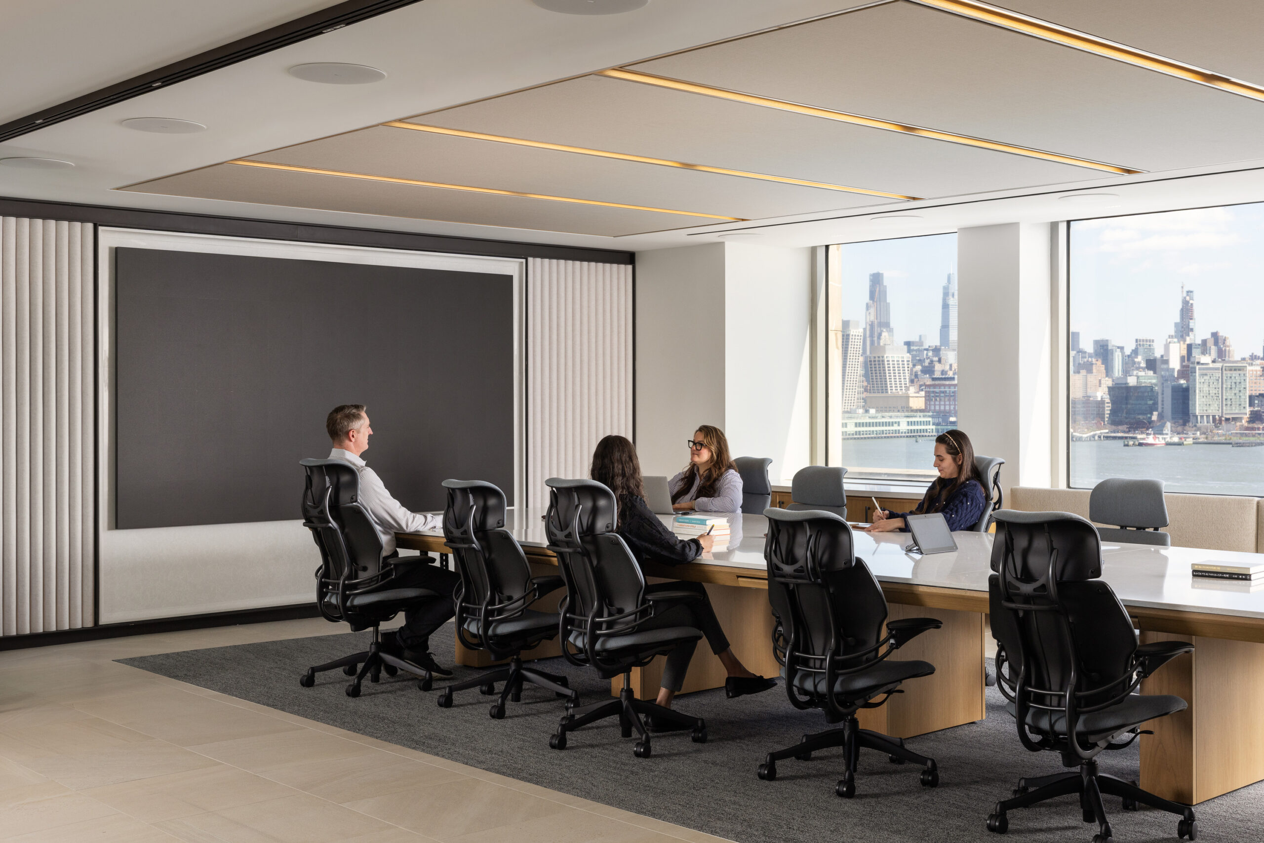 Modern conference room featuring a sleek, rectangular table surrounded by ergonomic black office chairs. A prominent wall-mounted screen offers a minimalist backdrop. Expansive windows reveal a stunning city skyline, flooding the space with natural light and enhancing the professional atmosphere. Neutral tones create a sophisticated ambiance.