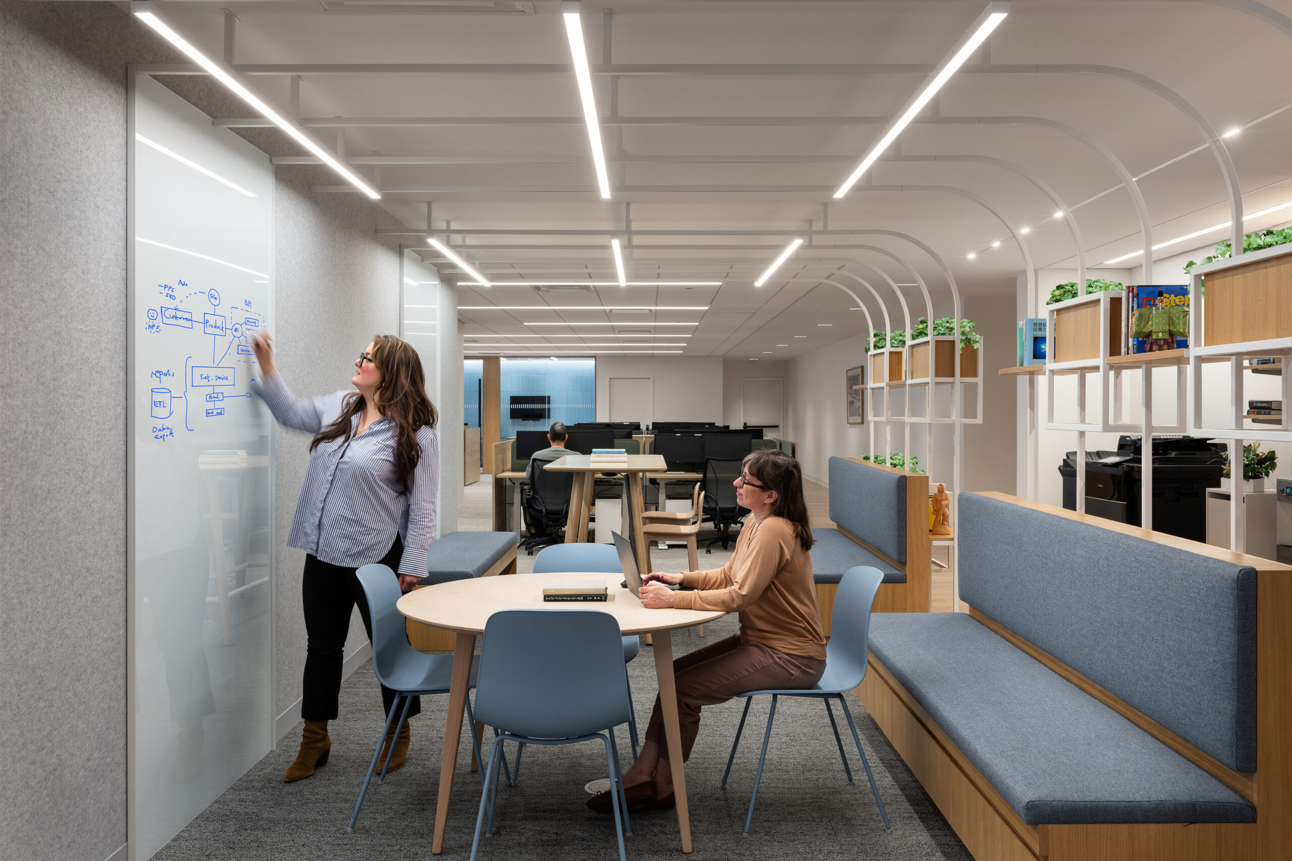 A modern office environment featuring collaborative workspaces. A woman writes on a whiteboard while another sits at a round table, both surrounded by sleek, minimalist furniture. Natural light floods the space, accentuated by innovative overhead lighting, with greenery adorning the shelves, promoting a refreshing and productive atmosphere.