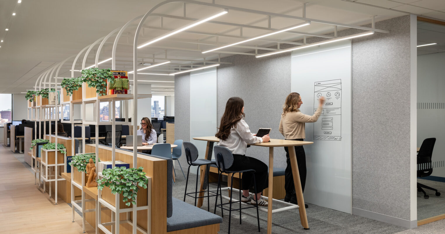 A modern office space featuring collaborative work zones. Two women engage in creative brainstorming at a standing-height table with minimalist design, surrounded by potted greenery. Clear glass partitions and a soft color palette enhance the open feel, while ergonomic seating and organized shelving promote productivity and comfort.