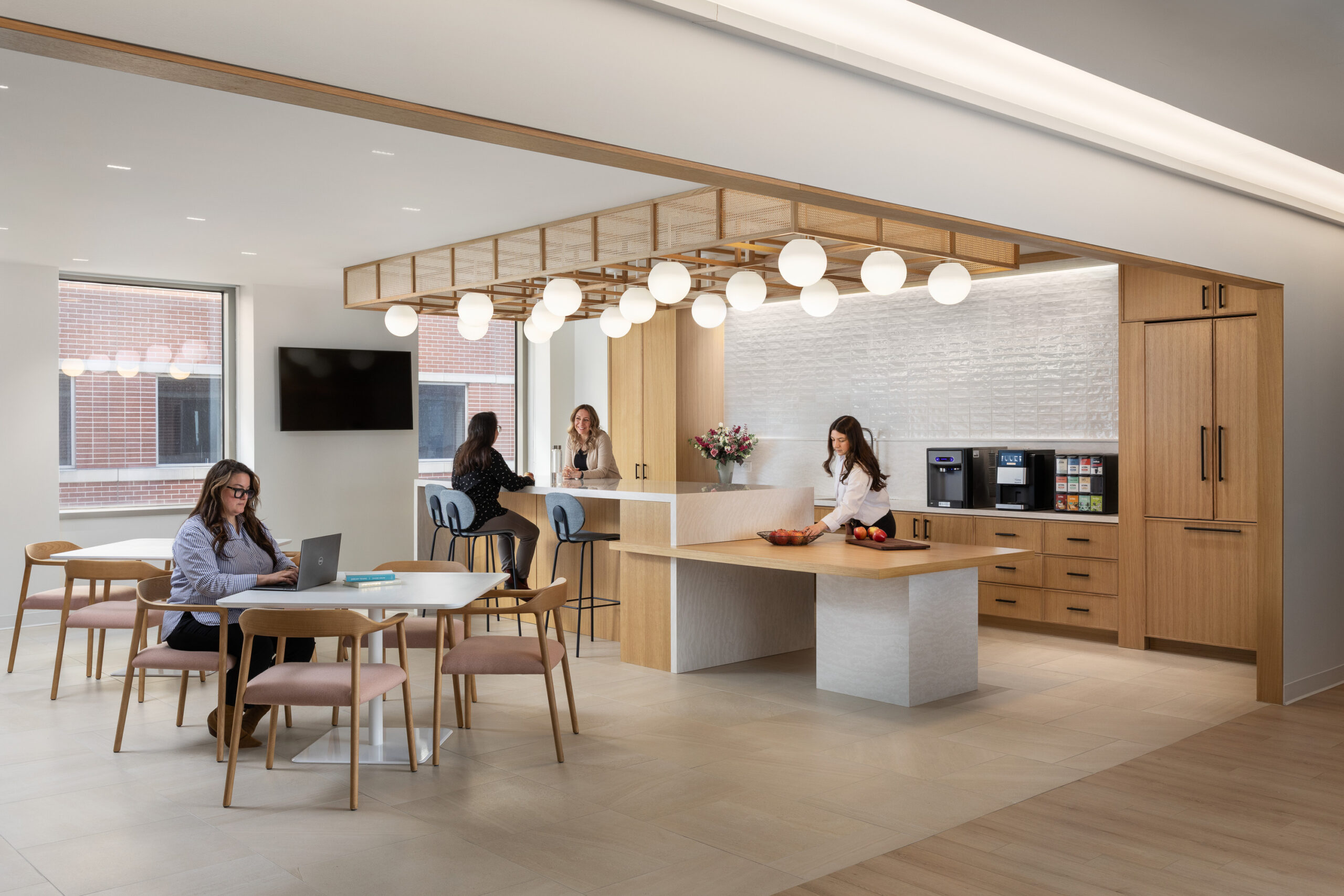 Bright, contemporary office kitchen featuring a sleek island with a white stone countertop. Elegant wooden cabinetry and a textured wall enhance texture and warmth. Modern pendant lights create a welcoming atmosphere, while workers engage in collaborative tasks at a dining table, promoting a functional and inviting workspace.