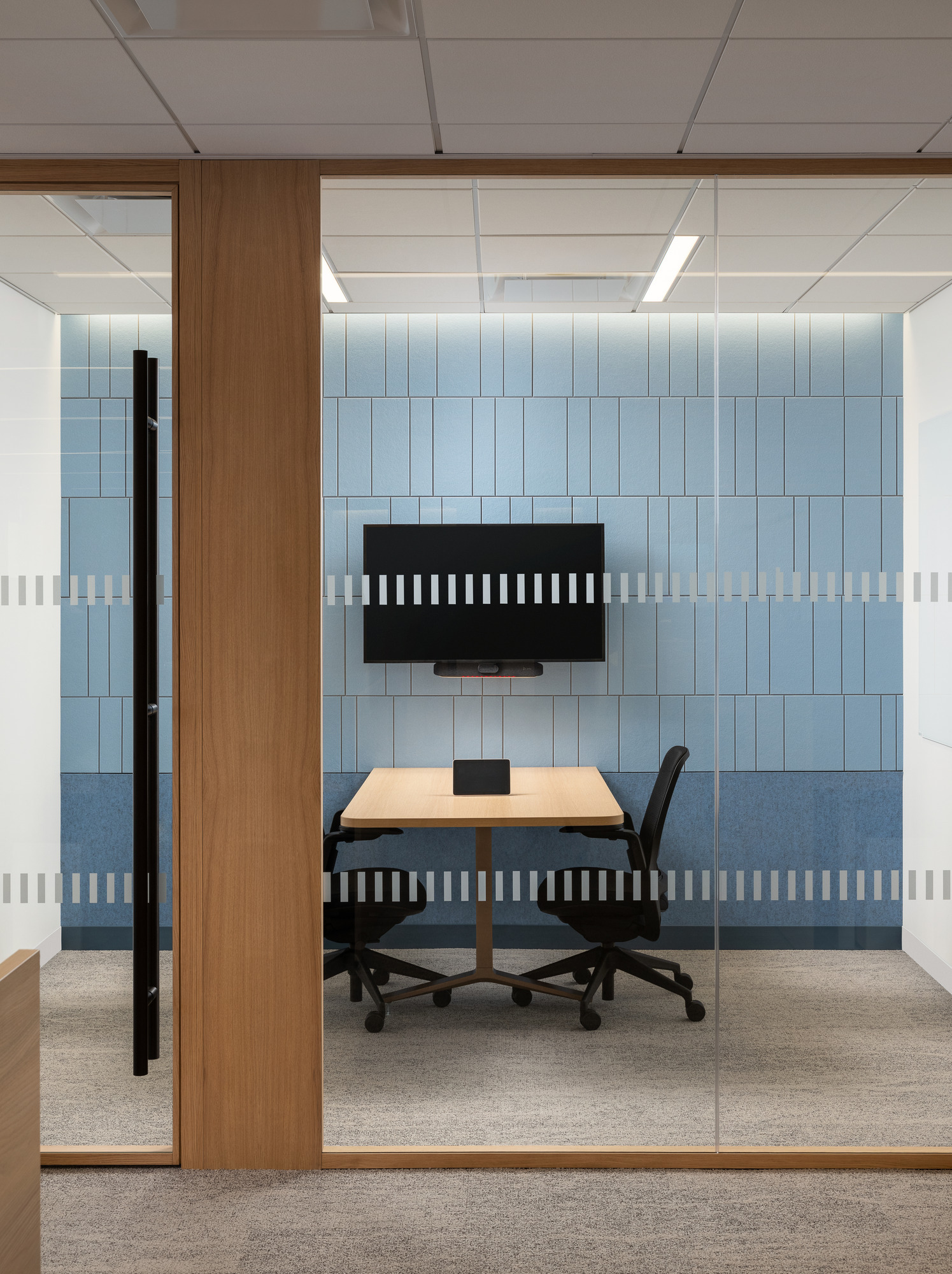 Brightly lit conference room featuring a linear blue wall design complemented by light wood accents. A sleek wooden table is paired with an ergonomic black chair, while a wall-mounted screen provides modern technological integration. Transparent glass walls enhance openness, promoting collaboration in a contemporary office environment.